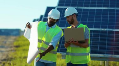 Mühendislerle birlikte Afrika kökenli Amerikalı bir adam güneş çiftliğinde özel bir üniformayla proje planının arkasında duruyor. Güneşli pillerin kurulumunu tartışıyorlar. Elektrik yavaş çekim.