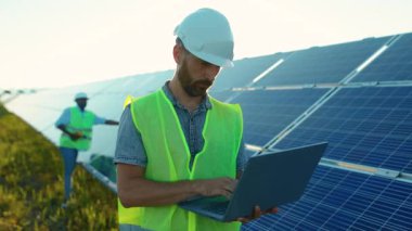 Genç adam dizüstü bilgisayar kullanıyor. Güneş pillerinin yakınındaki kameraya bak. Günışığı teknolojisinde yenilenebilir enerji işçisi.