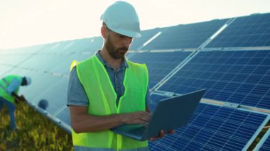 Çekici genç adam dizüstü bilgisayar kullanıyor. Güneş pillerinin yakınındaki kameraya bak. Günışığı teknolojisinde yenilenebilir enerji işçisi.