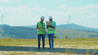 Güneşli bir günde, iki genç mühendis tarlalarda yel değirmenlerinin yanında dizüstü bilgisayar kullanıyor. İş birliği, rüzgar enerjisi tarım teknisyenleri yavaş çekim konuşuyor.