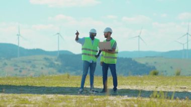 Ciddi iki genç mühendis, saha çalışmalarında yel değirmenlerinin yanında dizüstü bilgisayar standı kullanıyor. Yavaş çekim konuşan rüzgar enerjili tarım teknisyenleri işbirliği hakkında tartışıyorlar.
