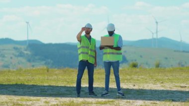 İki genç mühendis, iş yerlerinde yel değirmenlerinin yanında dizüstü bilgisayar standı kullanırlar.