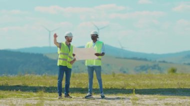 Mühendisler proje planıyla birlikte sahada yel değirmenlerinin yanında durup ekip çalışması yapıyorlar. Rüzgâr enerjili tarım teknisyenleri yavaş çekim konuşuyorlar.