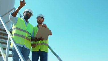 Yavaş çekim. Üniformalı iki mühendis dizüstü bilgisayar kullanıyor. Rüzgar türbinleri, ekolojik enerji endüstrisi.