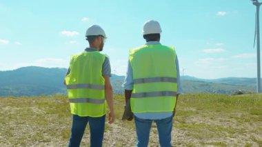 Yakın çekim. İki mühendis, tarlalarda yel değirmenlerinin yanında duruyor. Ekip, iş birliği, rüzgar enerjisi tarım teknisyenleri, ağır çekim konuşuyor.