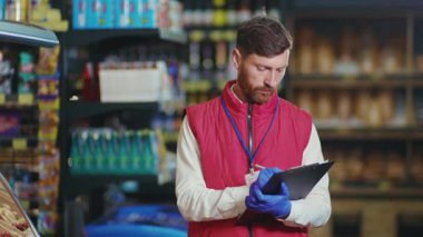 Kapalı gişe genç satıcı süpermarkette deftere bir şeyler yazıyor. Kameraya bak. Ciddi sebze işi. Güzel, taze, perakende satış mağazası.