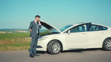 Mutsuz işadamı telefonda konuşuyor. Bozuk arabanın yanında oturuyor. Kaputu açtı.