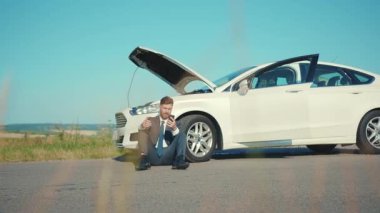 Sinirli işadamı, bozuk arabanın yanında oturan telefonu kullanıyor. Kaputu açtı. Stres sorununu düzeltmeye yardım ediyor.