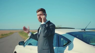 İş elbiseli kızgın adam, yolda akıllı telefonla konuşurken arabanın yanında duruyor ve güneş ışığında iş görüşmesi yapıyor. Yakışıklı telefon işi ağır çekim.