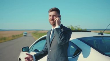 İş elbisesi içinde gülümseyen adam yolda akıllı telefonda konuşurken arabanın yanında duruyor ve güneş ışığında iş görüşmesi yapıyor. Yakışıklı telefon işi ağır çekim.