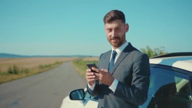 Takım elbiseli mutlu adam arabanın yanında duruyor. Gün ışığında akıllı telefon kullan. Gün ışığında konuş.