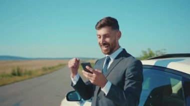 İş elbiseli çekici genç adam arabanın yanında duruyor. Gün ışığında yol konuşmasında akıllı telefon kullanıyor. Yakışıklı telefon işi ağır çekim.