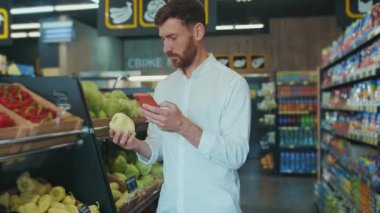 Süpermarkette duran yakışıklı genç adam telefondaki alışveriş listesine bakıyor. Sebze müşterileri seçiyor. İş yemekleri dükkânı.