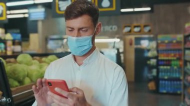 NEW YORK - 19 Nisan 2020: Süpermarkette maskeli genç adam telefondaki alışveriş listesine bakıyor sebze müşterileri için gıda mağazası yetişkin pazarı erkek perakende yavaş çekim