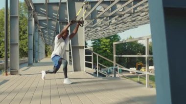 NEW YORK - 5 Eylül 2020: Güneşli Afrika kökenli Amerikalı genç adam, Trx fitness halatıyla dışarıda vücut egzersizleri yapıyor, stadyum kas antrenmanları, spor kıyafetleri spor antrenmanları, yavaş çekim egzersizleri.