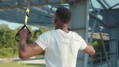 NEW YORK - 5 Eylül 2020: Güneş ışığında yakışıklı Afro-Amerikalı adam, Trx fitness halatıyla dışarıda vücut egzersizleri yapıyor stadyum kaslı spor kıyafetleri spor antrenmanı ağır çekim
