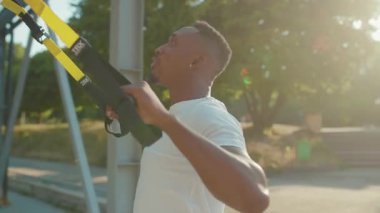 NEW YORK - 5 Eylül 2020: Yüzünüzü kapatın yakışıklı Afro-Amerikan adam trx fitness ipi ile çalışıyor açık hava vücut egzersizleri stadyumda güneşli bir günde kas antrenmanı spor kıyafetleri spor antrenmanı yavaş çekim