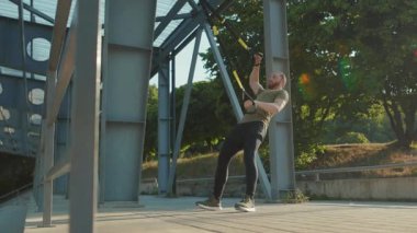 NEW YORK - 5 Eylül 2020: Trx fitness halatıyla çalışan yakışıklı genç adam güneşli bir günde stadyumda vücut egzersizleri yapıyor.