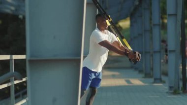 NEW YORK - 5 Eylül 2020: Güçlü, yakışıklı, Afro-Amerikan adam, Trx fitness halatıyla dışarıda vücut egzersizleri yapıyor, stadyum kaslı spor kıyafetleri spor kıyafetleri spor antrenmanı ağır çekimde.