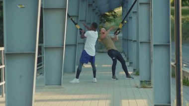 NEW YORK - 5 Eylül 2020: Güneşli bir günde yakışıklı iki adam, trx fitness halatıyla dışarıda vücut egzersizleri yaparak stadyumda Afrika kökenli Amerikan spor giyim sporu antrenmanlarını yavaş çekimde yapıyorlar.