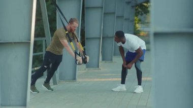 NEW YORK - 5 Eylül 2020: Afrikalı Amerikalı genç adam, spor giyim sporları ağır çekimde stadyum kaslı spor antrenmanlarında trx fitness halatıyla vücut egzersizleri yapan birini motive etti