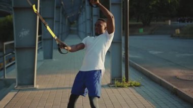 NEW YORK - 5 Eylül 2020: Trx fitness halatıyla çalışan ciddi yakışıklı Afro-Amerikalı adam stadyum kaslı spor kıyafetleri spor antrenmanı ağır çekimde vücut egzersizleri yapıyor.