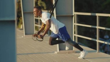 NEW YORK - 5 Eylül 2020: Güçlü, yakışıklı, Afro-Amerikan adam, Trx fitness halatıyla dışarıda vücut egzersizleri yapıyor, stadyum kaslı spor kıyafetleri spor kıyafetleri spor antrenmanı ağır çekimde.