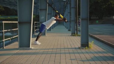 NEW YORK - 5 Eylül 2020: Güneş ışığında yakışıklı Afrikalı Amerikalı adam, Trx fitness halatıyla dışarıda vücut egzersizleri yapıyor stadyum kaslı spor kıyafetleri spor kıyafetleri spor antrenmanı ağır çekim
