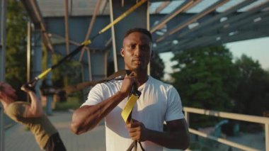 Ciddi Afrikalı Amerikalı adam trx fitness halatıyla dolaştıktan sonra kameraya bakıyor. Stadyumdaki kas sporları spor kıyafetleri spor salonunda vücut egzersizleri. Yavaş çekim yakın çekim.