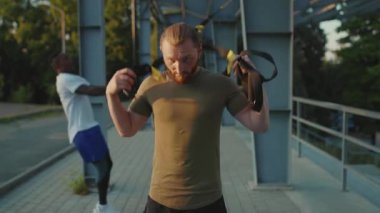 Ciddi yakışıklı adam, trx fitness halatıyla dolaştıktan sonra kameraya bak. Stadyumdaki spor kıyafetleri spor salonunda vücut egzersizleri. Yavaş çekimde spor portresi kapat.
