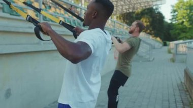 Yaklaş genç adam. Trx fitness halatıyla dışarıda vücut egzersizi yapıyor. Stadyum kaslı spor kıyafetleri, spor kıyafetleri, çekici göğüs egzersizleri, ağır çekim.