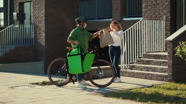 Güvenlik kaskı ve sağlık maskesi takmış genç bir kurye müşteriye bisiklet çekici bir kadın ile kese kağıdıyla yemek getiriyor dışarıda durup Coronavirus 'un daha az yavaş çekim teması kurmasını söylüyor.