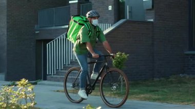 Dışarıda güvenlik kaskı ve tıbbi maske takmış genç bir kurye bisiklet siparişi Coronavirus ile yemek getiriyor.