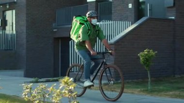 Sokakta güvenlik kaskı ve sağlık maskesi takmış genç bir kurye bisiklet siparişi Coronavirus ile yemek getiriyor.