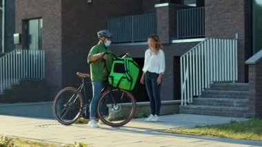 Güneş ışığı kuryesi kasklı ve tıbbi maskeli bir kurye müşteriye bisiklet çekici bir kadın eşliğinde kese kağıdıyla yemek getiriyor dışarıda durup Coronavirus 'un daha az yavaş çekim teması kurmasını söylüyor.