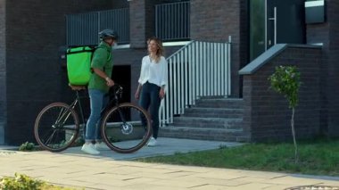 Genç adam kuryesini güvenlik kaskı ve tıbbi maskeyle kapatın. Müşteriye bisiklet çekici bir kadınla kese kağıdıyla yemek dağıtın. Dışarıda durun.