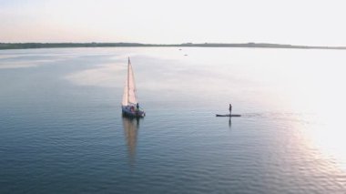 Birdseye sinematik huzurlu nehir manzarası, yelkenli beyaz yat ve sup sörfü yapan turistleri çekiyor. Tatil, yaz aktiviteleri ve eğlence kavramı.