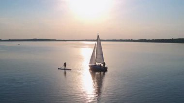 Sinema günbatımında durgun nehir suyunda yüzen beyaz bir yat. Sup-surf yapmaktan hoşlanan bir turist. Hava görüntüsü. Doğal konsept.