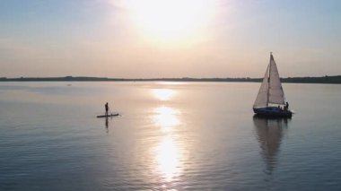 Harika bir göl. Su aktiviteleri. Bir grup genç turist beyaz yatta yelken açarken, bir adam da sup sörfünün tadını çıkarıyor. İHA uçuşunda sahne doğası.