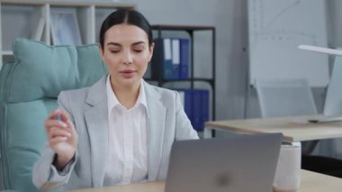 Gri takım elbiseli hoş görünümlü genç bir kadının görüntüsü dizüstü bilgisayarın başında oturup konuşması, video görüşmesi sırasında notlar konuşması. Resmi kıyafetli gülümseyen iş kadını portresi. İçeride. Ofis. İş yeri. Çevrimiçi toplantı