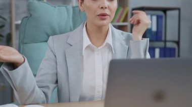 Gri takım elbiseli beyaz iş kadınının webcam 'e bakması, internet üzerinden iş görüşmesi yapması, müşteriyle konuşması, mesafesi, vesikalık fotoğrafı. Masada oturan ve notlar alan güzel bir genç kadın.