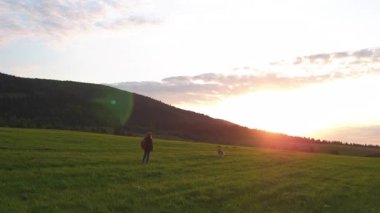 Tepelerde duran ve gökyüzünde gün batımını seyreden bir kadın. Güzel dağ manzarasında köpeğiyle oynayan mutlu beyaz kız..