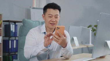 İş yerinde oturup akıllı telefon ekranına bakan, ofiste sohbet eden neşeli görünümlü, hoş görünümlü, yetişkin Asyalı bir satıcının yatay görüntüleri. İşte beyaz gömlekli gülümseyen Çinli adam. İçeride. Görev