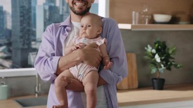 Yakışıklı beyaz bir babanın, güzel bir kızı elinde tutarken gülüşünün görüntüsü. Mutlu baba ve kızının portresi. Pembe elbiseli sevimli çocuk kameraya bakıyor. Modern iç mekan. Mutfak.