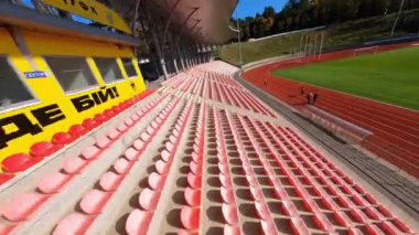 FPV, güneşli bir günde büyük çağdaş stadyumun üzerinde manevra yapabilir. Renkli tribün koltuklar, çimen futbol sahası ve sporcular eğitimi.