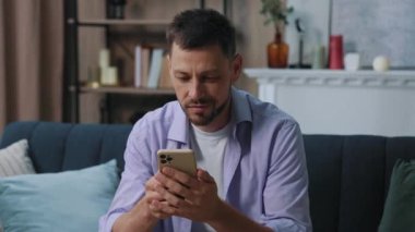 Çekici, gülümseyen, mutlu genç adam evdeki koltukta oturan telefonu kullanıyor. İletişim işadamı sosyal medyada sohbet ediyor. Kapatın. Portre. Yavaş çekim