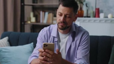 Yakışıklı genç adam evdeki kanepede otururken telefonla gülümsüyor. İletişim işadamı sosyal medyada sohbet ediyor. Kapatın. Portre. Yavaş çekim