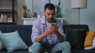 Acaba gülümseyen mutlu genç adam evdeki koltukta otururken telefon kullanıyor mudur? Portre. İletişim işadamı sosyal medyada sohbet ediyor. Kapatın. Portre. Yavaş çekim