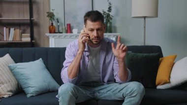 Çekici genç adam kanepede oturuyor ve cep telefonuyla konuşuyor. Modern daire. Ciddi sakallı adam. Yavaş çekim