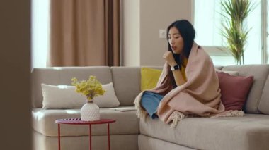 Kanepede battaniyeyle oturan Asyalı genç bir kadının portresi evde kendini kötü hissediyor. Salgın. Coronavirus. Enfeksiyon kapmış hastalık stres altında. Hastalık. Yavaş çekim